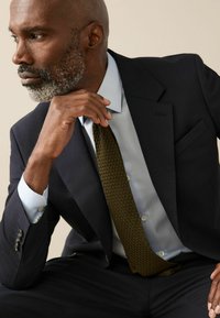 Dunkler Anzugjacke über einem hellblauen Hemd, strukturierte olivgrüne Krawatte, Hände ruhen am Kinn. Dezenter Gesichtsbart und Hautstruktur sichtbar.