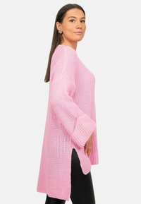 Overmaatse roze gebreide trui met brede mouwen, zijsplitten en een textuurpatroon. Draag je over zwarte leggings.