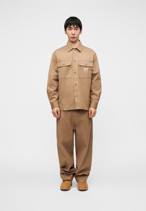 Tan cotton shirt with two front pockets, button closure, and a logo. Paired with matching loose-fit pants and tan footwear.