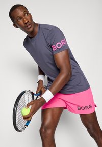 Vīriešu tenisa tērps sastāv no pelēka īsām piedurknēm krekla ar rozā „BORG” uz piedurknes un rozā šortiem ar „BORG” logo. Spēlētājs tur tenisistu raketi un bumbu.
