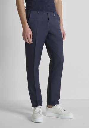 Man draagt donkerblauwe op maat gemaakte broek en witte sneakers, staand tegen een effen lichte achtergrond.