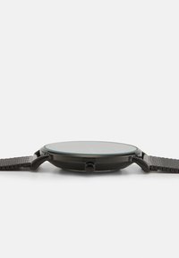 Montre noire avec un cadran rond et plat, bracelet en maille métallique et surface en verre lisse. Présente un design minimaliste et une couronne latérale pour les ajustements.