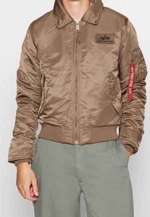 Brun bomberjakke med lynlås foran, ribstrikkede ærmeafslutninger og kant, sidelommer, Alpha Industries-logo på lap, og rød "Remove Before Flight"-mærkat på ærmelommen.