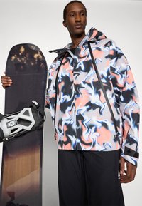 Giacca colorata con motivo astratto, cappuccio, dettagli neri e cerniere. Include uno snowboard scuro e attacchi grigi. Tessuto strutturato.