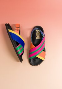 Paire de sandales plates colorées avec des brides croisées en rose, vert, bleu, jaune et orange sur un fond dégradé pêche.