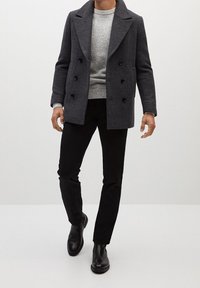 Manteau en laine gris à double boutonnage, porté sur un pull gris clair. Associé à un pantalon noir slim et des bottines noires.