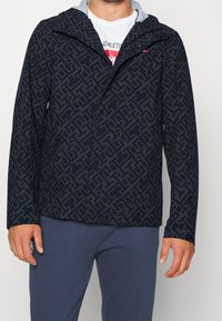 Námořnický větrovka s geometrickým vzorem, kapucí a šedou podšívkou. Tkanina je hladká, s volným střihem a mírně elastickými manžetami.