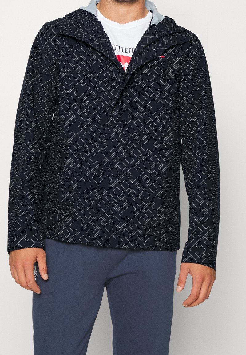 Námořnický větrovka s geometrickým vzorem, kapucí a šedou podšívkou. Tkanina je hladká, s volným střihem a mírně elastickými manžetami.