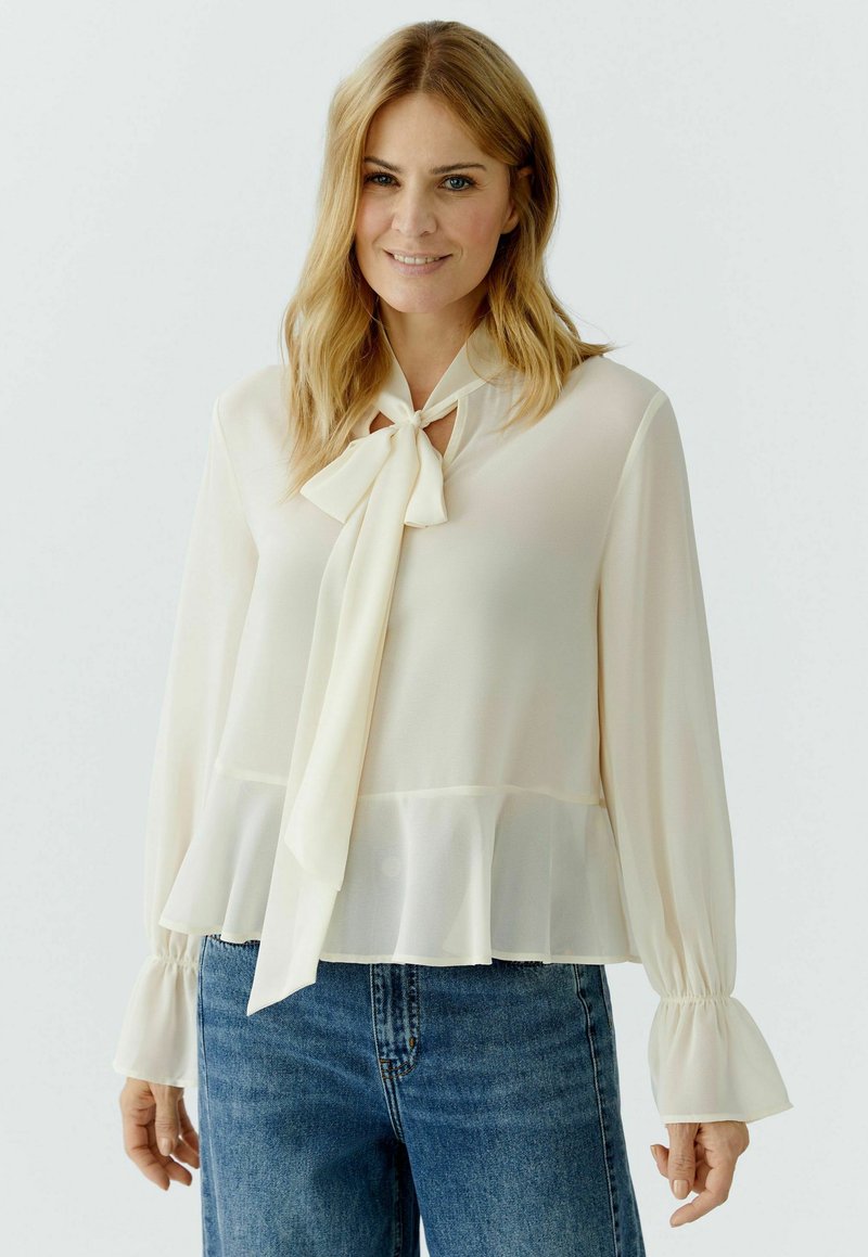 Woman with blonde hair wearing a cream long-sleeve blouse with a bow tie and ruffled cuffs paired with blue denim jeans.