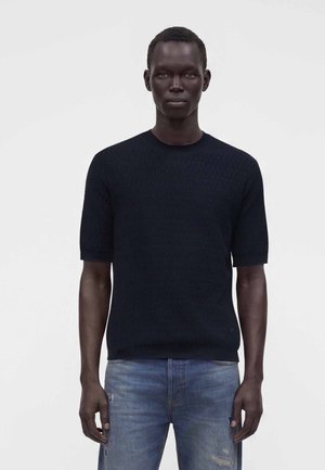 Homme à la peau foncée portant un pull noir texturé à manches courtes et un jean bleu clair déchiré, debout devant un fond clair uni.