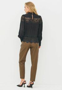 Czarna bluzka z koronkowymi akcentami i wysokim kołnierzem, w połączeniu z oliwkowozielonymi spodniami o tapered fit oraz czarnymi szpilkami. Posiada zapięcie na guziki z tyłu.