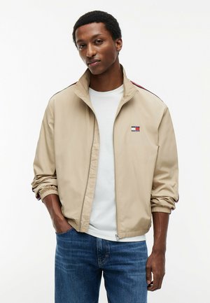 Mand iført beige lynlåstrøje over hvid skjorte med hænderne i lommerne, stående mod hvid baggrund.