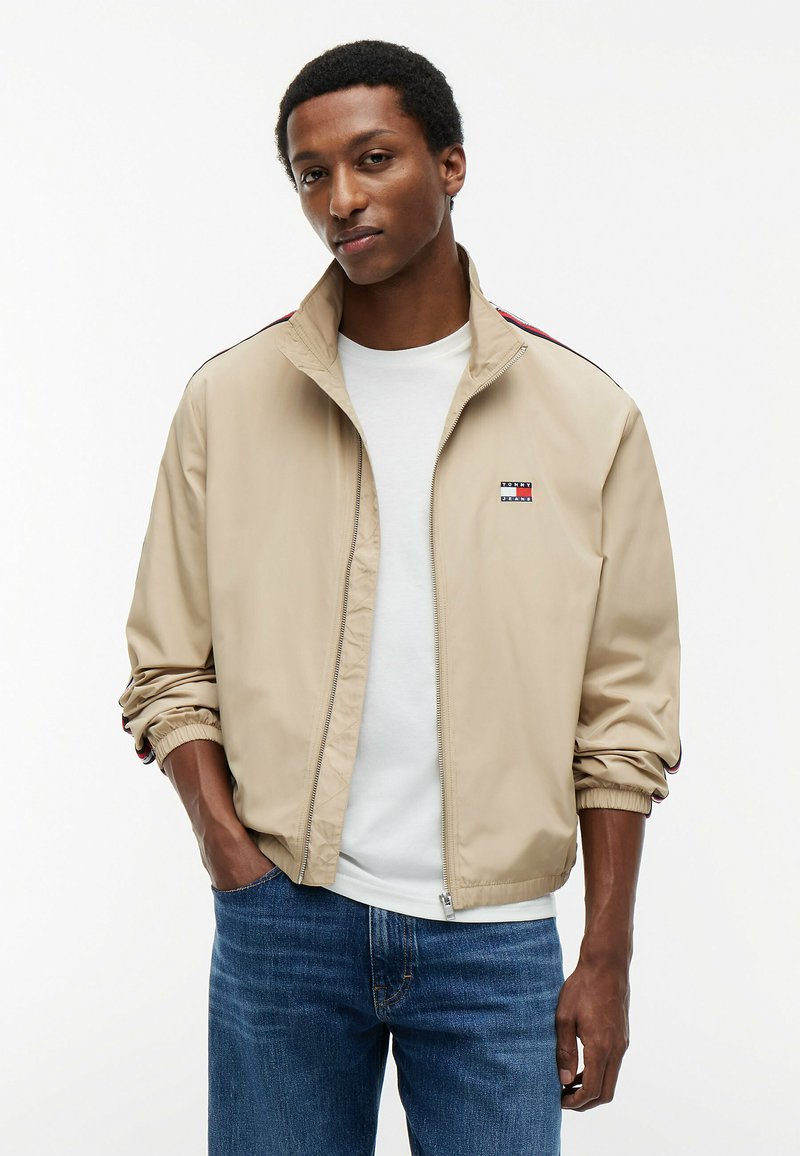 Homme portant une veste beige zippée sur une chemise blanche, les mains dans les poches, debout devant un fond blanc.