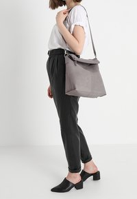 Šedá ramenní taška s překlopným víkem, hladkým povrchem, stříbrnými kovovými detaily a jedním nastavitelným popruhem. Nosená s černými zúženými kalhotami.