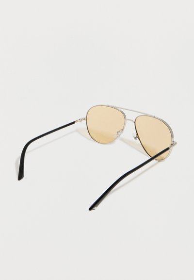 Ασημένια γυαλιά ηλίου αεροπόρου με ανοιχτομπεζ φακούς και μαύρα κυρτά μπράτσα που ακουμπούν σε λευκή επιφάνεια.