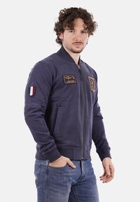 Giacca bomber navy con zip, polsini a costine, due tasche, toppe sul petto e sulla manica, e una texture liscia.