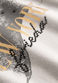 Lichtgrijze stof met een zwart-witte grafische print en gouden tekst. Versierd met zwarte rhinestones in een gebogen patroon.