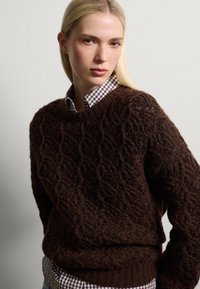 Brauner Strickpullover mit strukturiertem, gemustertem Design. Gerippter Halsausschnitt und Bündchen. Wurde über einem karierten Hemd mit einem weißen und braunen Muster getragen.
