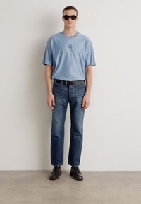 Light blue short-sleeve t-shirt with a logo, paired with dark blue jeans and black shoes. Model wears sunglasses, standing against a plain backdrop.