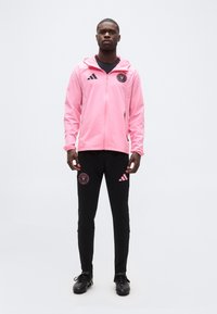 Άνδρας που φοράει ροζ αθλητικό μπουφάν με φερμουάρ και μαύρο παντελόνι προπόνησης με λογότυπα, στέκεται μπροστά σε απλό λευκό φόντο.