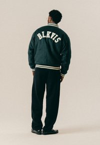 Lühikeste lokkidega mees, kes kannab prille ja musta ülikoolijakki, millel on tekst "BLKVIS", musti pükse ja musti jalanõusid, seismas seljaga ees.