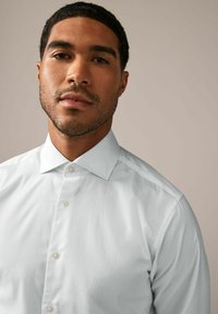 Hvid skjorte med knapper, der har en glat tekstur, en klassisk krave og en subtil glans, præget af synlige knapper nedad foran.