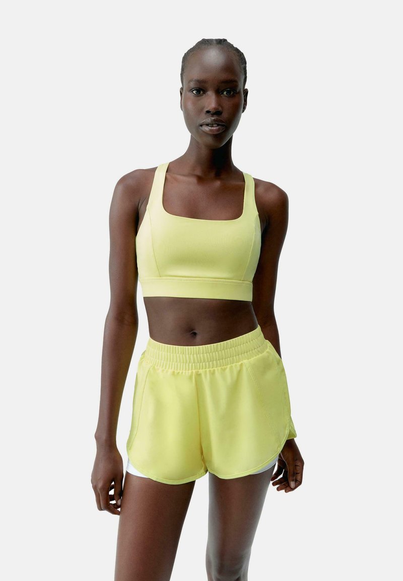 Geel sport-bh en bijpassende shorts van lichtgewicht materiaal. De top heeft een vierkante halslijn; de shorts hebben een elastische tailleband en zijsplitten.