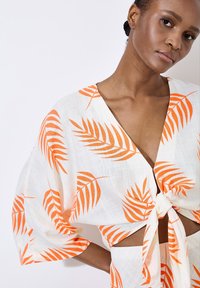 Hvid top med orange palmebladtryk, der har en dyb V-hals, brede ærmer og en frontbinding. Letvægtsstof med en tekstureret overflade.