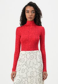 Rotes Langarmoberteil mit floralem Muster und hohem Kragen, kombiniert mit einem weißen Faltenrock, der schwarze Blumenumrisse aufweist.