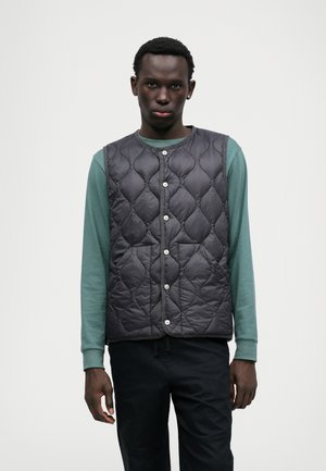 Κουβερτίνα χωρίς μανίκια σε σκούρο γκρι χρώμα με ασημί κουμπιά, φορεμένη από πάνω ενός τιραντό μπλουζάκι σε τιρκουάζ απόχρωση. Λείος υφασμάτινος υφή, ελαφρύ σχέδιο.