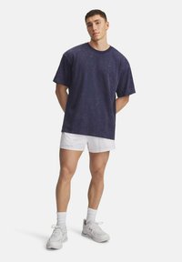 Junger Mann steht mit den Händen hinter dem Rücken, trägt ein dunkelblaues, gewaschenes T-Shirt, weiße Shorts, weiße Socken und weiße Sneaker.