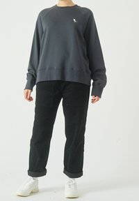 Felpa grigia scura con scollo tondo e maniche raglan, caratterizzata da un piccolo logo di un uccello bianco. Abbinata a pantaloni di velluto a coste neri e sneaker bianche.