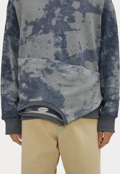 Gray-blue tie-dye sweatshirt with a unique angled hem and ribbed cuffs. Features a label at the bottom with text. Paired with beige pants.