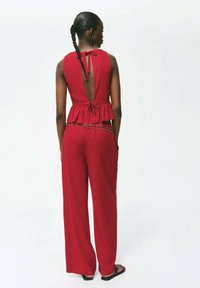 Rdež kombinezon z peplum pasom, odprtim hrbtom in povezovanjem. Ima široke hlače in akcent traku v pasu. Mehka tekstura tkanine.