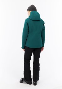 Mujer de espaldas vistiendo una chaqueta con capucha de color teal, pantalones de esquí negros, un gorro negro y botas de esquí negras sobre un fondo blanco.