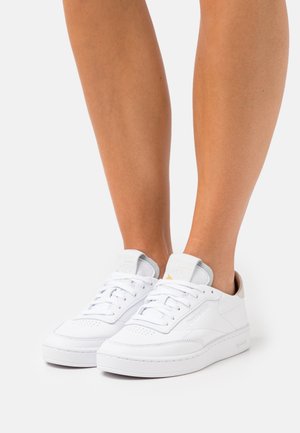 Jambes portant des baskets basses blanches à lacets blancs, avec un logo discret et des accents beiges au niveau du talon, sur un fond clair uni.