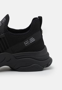 Černá sportovní obuv s prodyšným síťovinovým svrškem, texturovanou podrážkou, výraznými detaily a varovným nápisem na boku. Zaoblený design pro pohodlí.