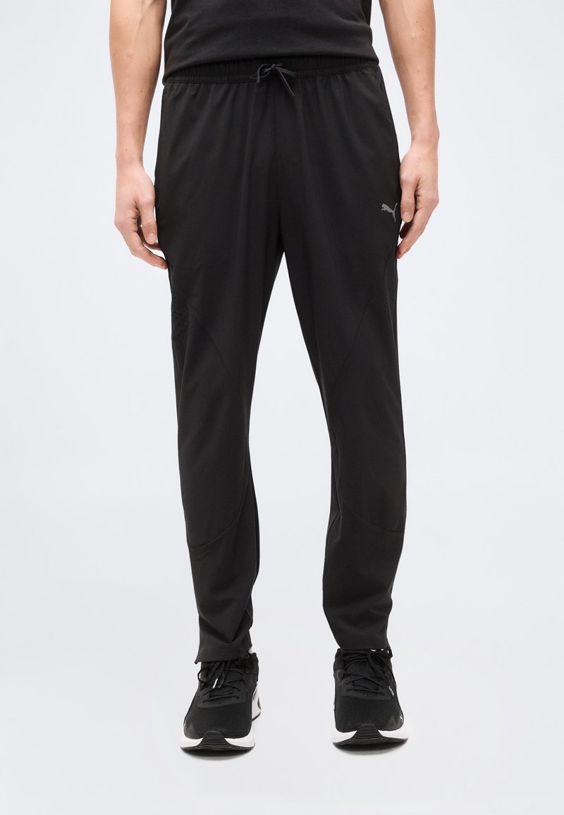 Άνδρας που φοράει μαύρο παντελόνι φόρμας Puma και μαύρα αθλητικά παπούτσια, στέκεται μπροστά σε ένα απλό ανοιχτόχρωμο φόντο.