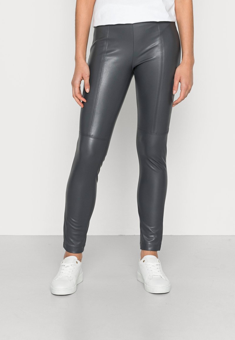 Grijze faux leren leggings met hoge taille, flatlock-naden, slanke pasvorm en een gladde textuur, gecombineerd met witte sneakers op een neutrale achtergrond.