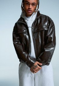 Bruine leren jas met een ritssluiting aan de voorkant, twee borstzakken en een ontspannen pasvorm, gedragen over een grijze hoodie en joggingbroek.