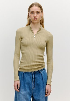 Beige geripptes Langarmoberteil mit hohem Kragen und halber Reißverschlussöffnung, kombiniert mit lockeren blauen Jeans. Glatte Textur.