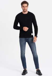 Schwarzer Strickpullover mit Rundhalsausschnitt, kombiniert mit blauen Slim-Fit-Jeans und schwarzen Sneakern. Auffälliges Etikett am Saum des Pullovers.