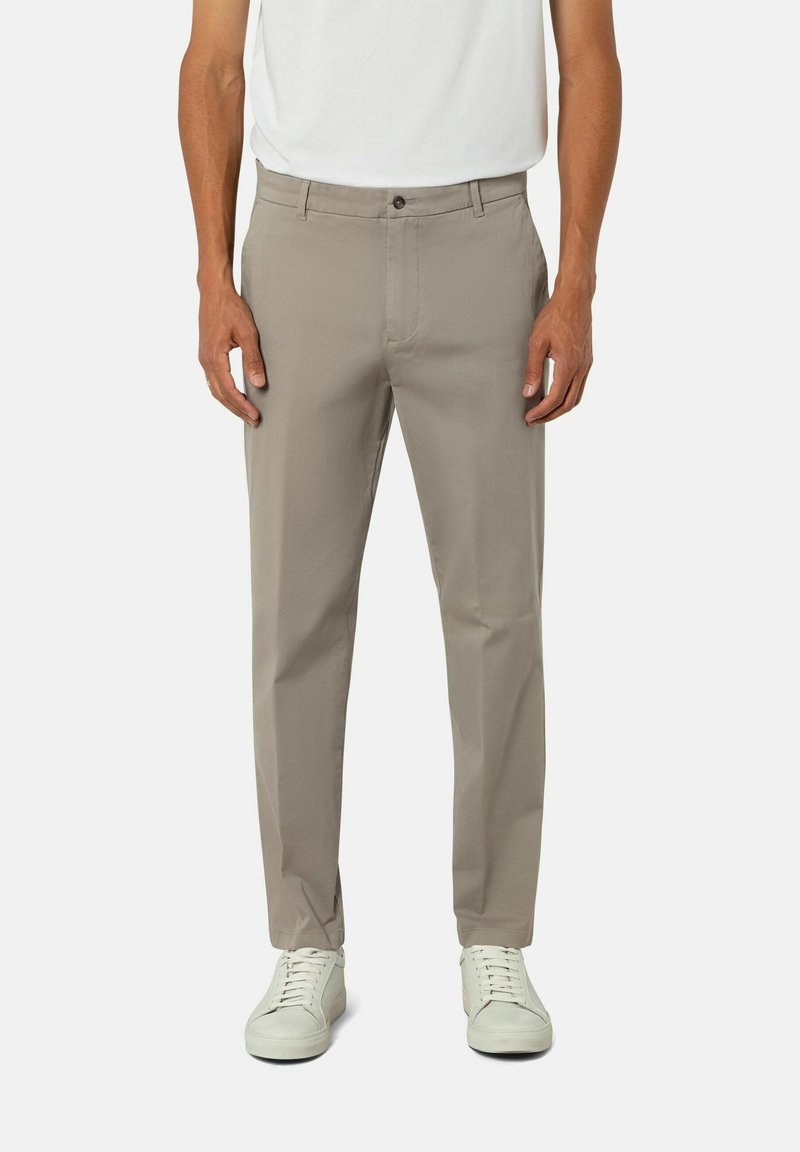 Beige Hose aus leichtem Stoff mit einem Knopfverschluss vorne, ausgestattet mit seitlichen Taschen und einem geraden Beinansatz. Getragen mit weißen Schuhen.