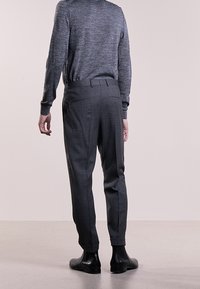 Hombre vestido con pantalones ajustados gris oscuro, suéter gris de manga larga y zapatos de cuero negros, de pie con la espalda hacia el frente contra un fondo liso.
