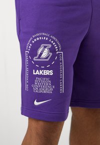 Lilla basketballshorts med et hvidt Nike-logo og Los Angeles Lakers-branding, herunder holdnavn og detaljer i fed skrift.