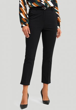 Vrouw draagt zwarte, op maat gemaakte enkelhoge broek met een smalle riem, gecombineerd met een meerkleurige blouse met geometrisch patroon en lange mouwen en zwarte hakken.