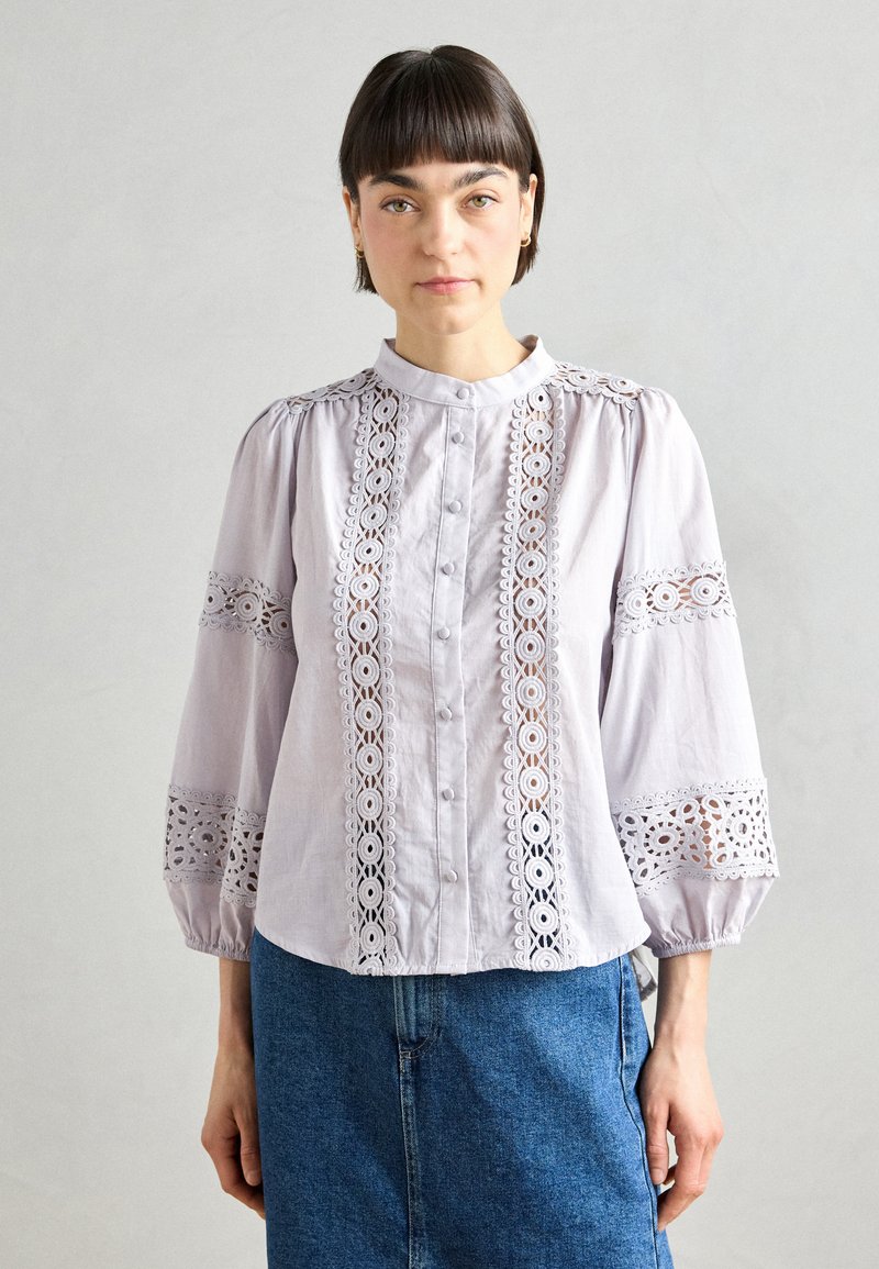 Gaiši lavandas blūze ar garām pūkainām piedurknēm, mežģīņu dekoratīviem elementiem priekšā un piedurknēs, kā arī pogu aizdari. Kombinēta ar džinsa svārkiem.