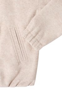 Manche et poche latérale d'une veste en polaire beige avec détails brodés et poignet élastiqué sur fond blanc.