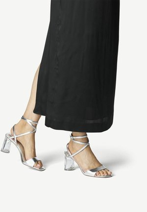 Feet wearing silver strappy high-heeled sandals with clear block heels, paired with a long black skirt.