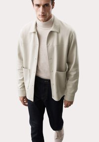 Chaqueta beige claro con cierre de botones, dos bolsillos laterales y cuello, combinada con un suéter de cuello alto beige y jeans de mezclilla oscuros.